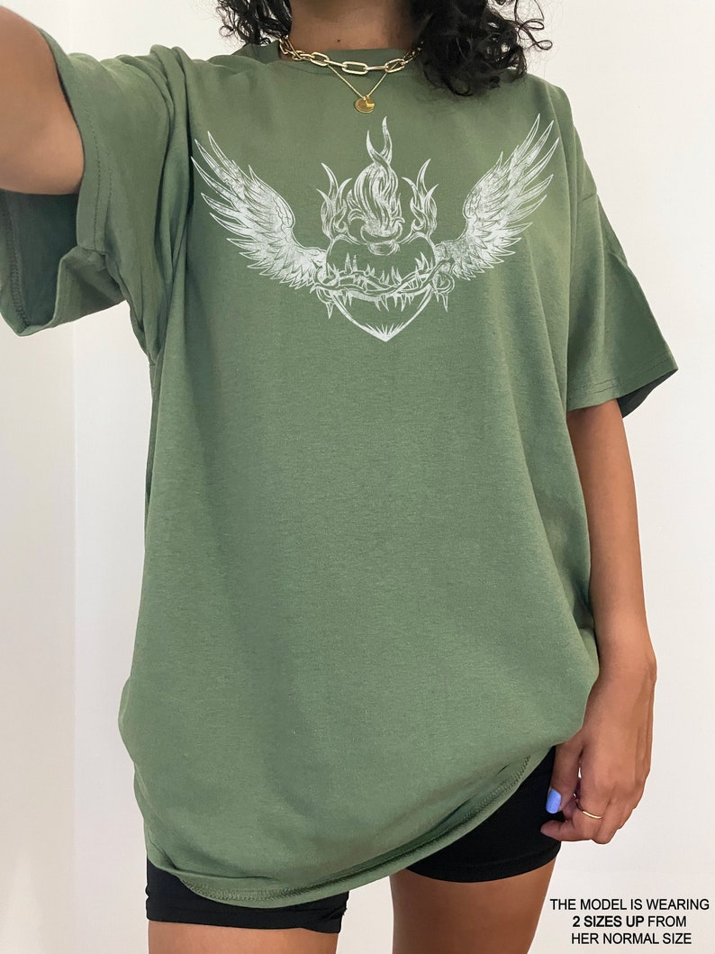 May include: Oversized green t-shirt featuring a white graphic of a heart with wings and flames. The heart is surrounded by a barbed wire design.