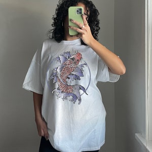May include: White oversized t-shirt with a graphic print of a red and purple koi fish swimming in blue water. The fish is surrounded by a circle and waves.
