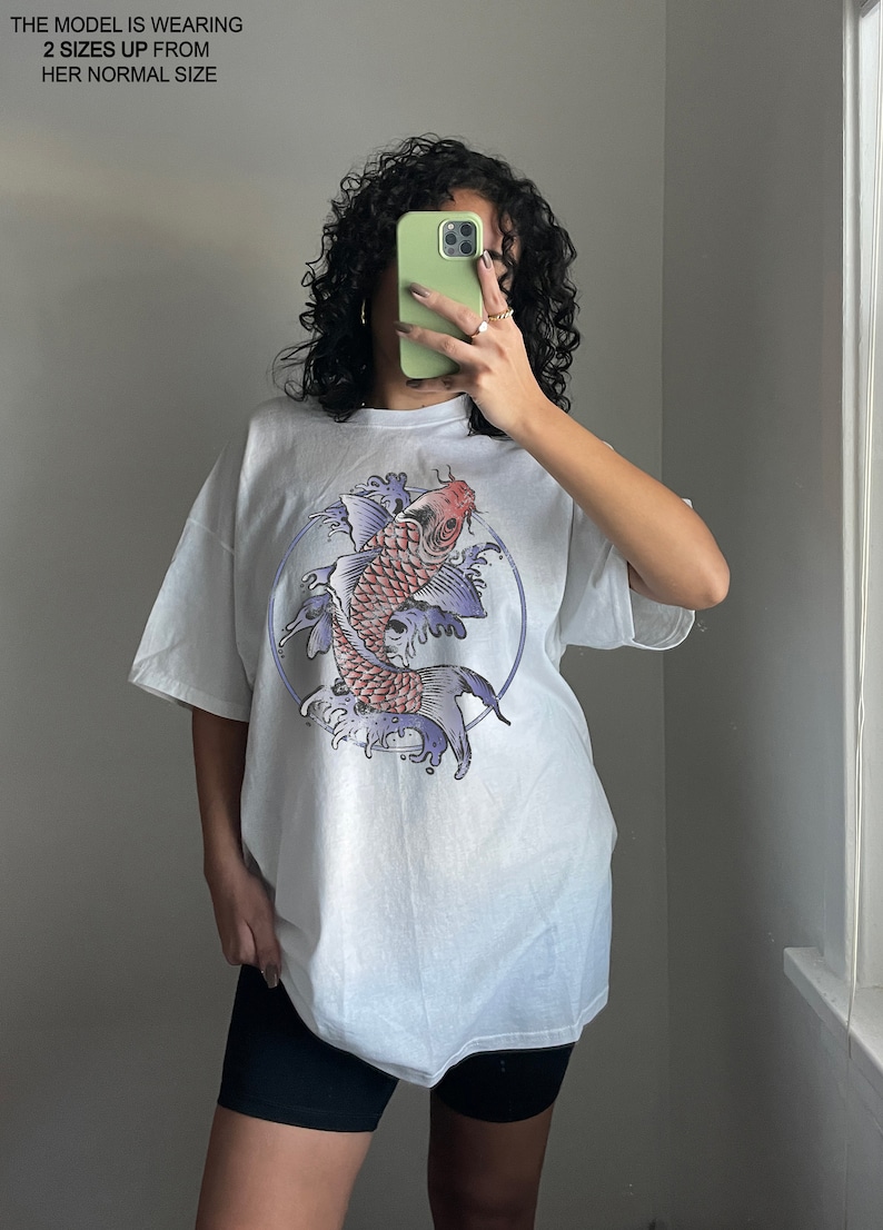 May include: White oversized t-shirt with a graphic print of a red and purple koi fish swimming in blue water. The fish is surrounded by a circle and waves.