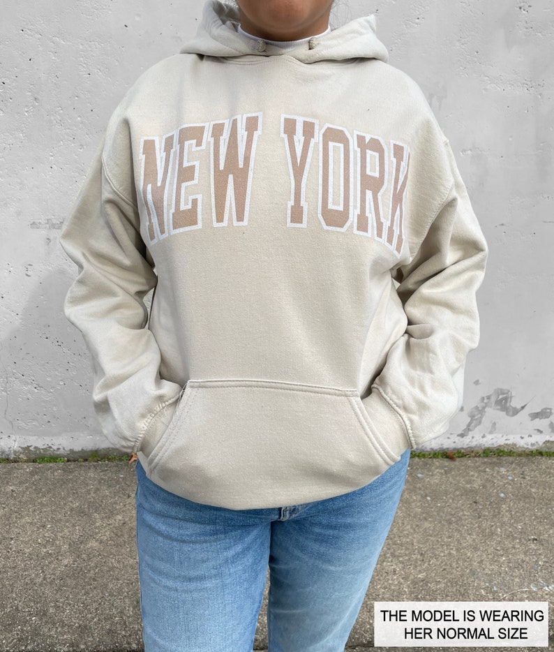 May include: A light beige hoodie with a large "NEW YORK" graphic printed in a darker beige color. The model is wearing a size that fits her normally.