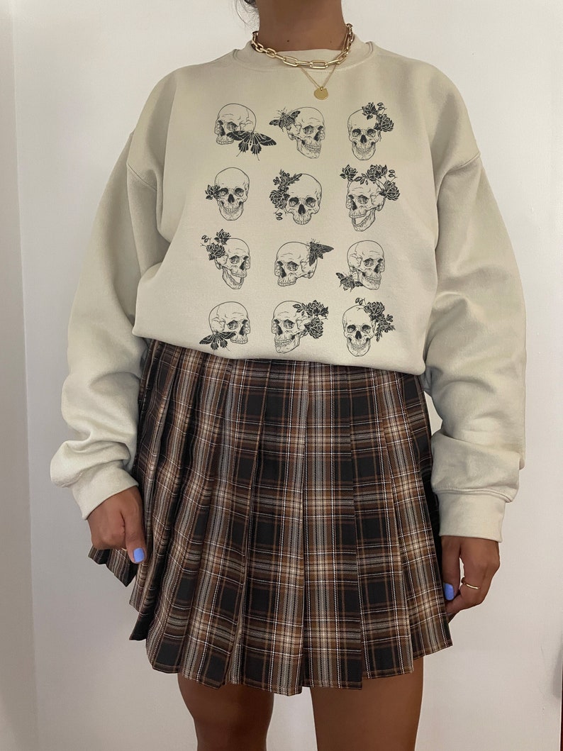 K&ouml;nnte beinhalten: Ein beiger Pullover mit einem grafischen Druck aus mehreren Sch&auml;deln, die mit floralen Akzenten und Schmetterlingen verziert sind. Der Pullover wird mit einem braunen und wei&szlig;en karierten Faltenrock kombiniert.