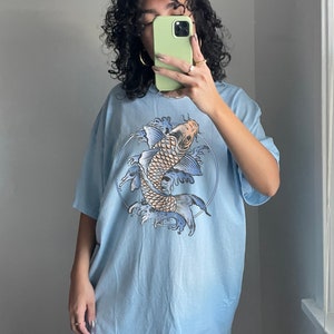May include: A light blue oversized t-shirt with a graphic print of a koi fish swimming in a circle of water. The fish is orange and white with blue and black details. The shirt is made of a soft, comfortable material.