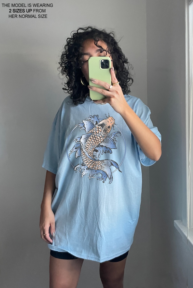 May include: A light blue oversized t-shirt with a graphic print of a koi fish swimming in a circle of water. The fish is orange and white with blue and black details. The shirt is made of a soft, comfortable material.