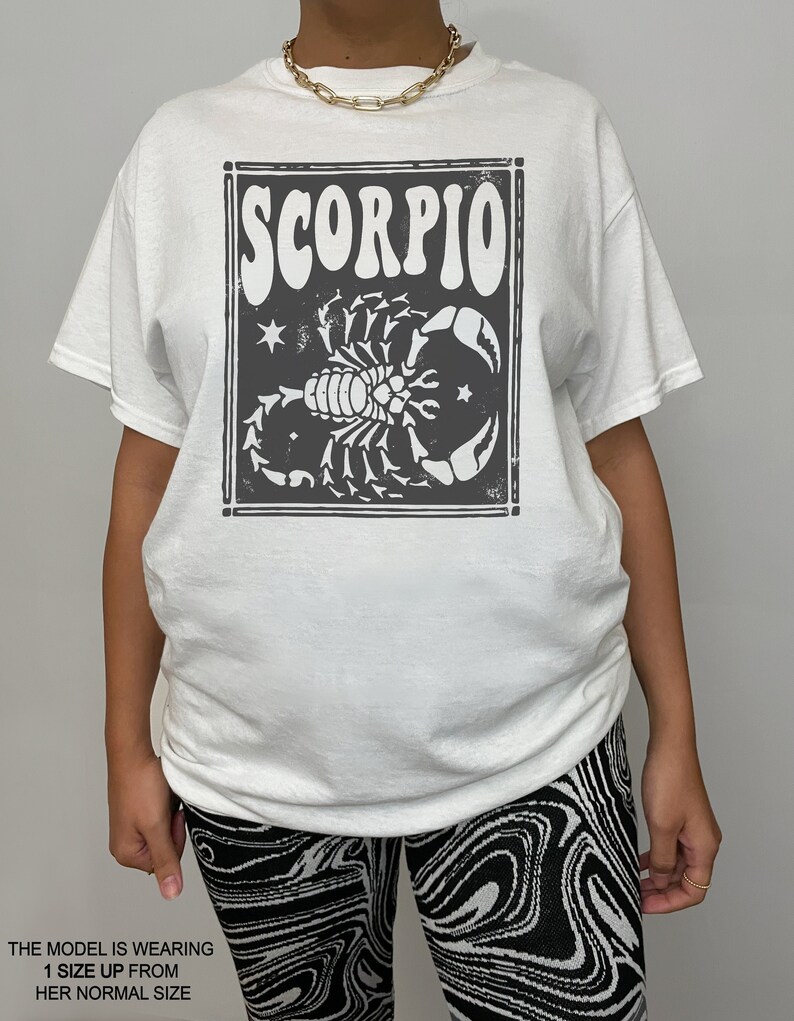 May include: White t-shirt with a black and white graphic print of a scorpion and the word "SCORPIO" in a retro font.