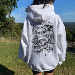 May include: White hooded sweatshirt with a black and white graphic print of multiple skulls on the back. The model is wearing the sweatshirt two sizes up from her normal size.