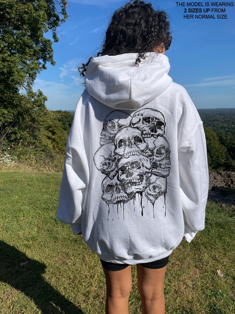 May include: White hooded sweatshirt with a black and white graphic print of multiple skulls on the back. The model is wearing the sweatshirt two sizes up from her normal size.