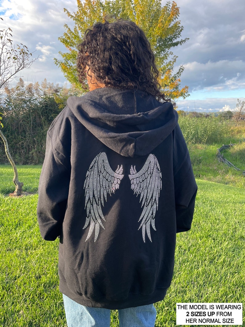 May include: Black hooded sweatshirt with a rhinestone design of angel wings on the back. The model is wearing the sweatshirt two sizes up from her normal size. "THE MODEL IS WEARING 2 SIZES UP FROM HER NORMAL SIZE"