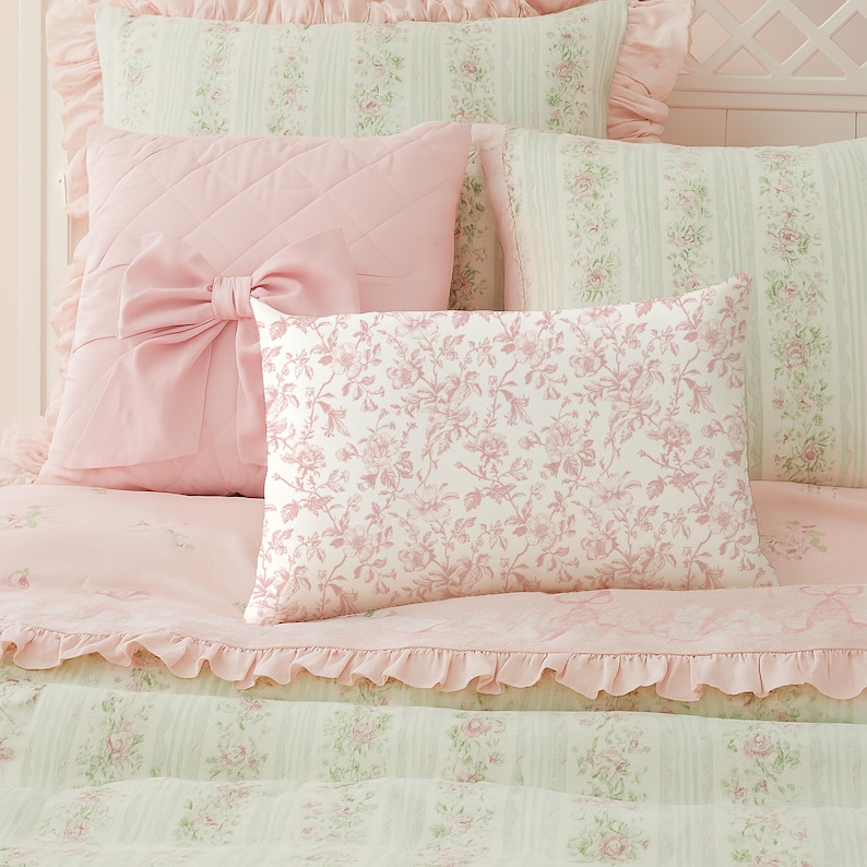 May include: A collection of pink and white floral bedding. The set includes a quilted pillow with a large bow, a rectangular pillow with a floral pattern, and several other pillows and a comforter with ruffled edges. The overall theme is romantic and feminine.