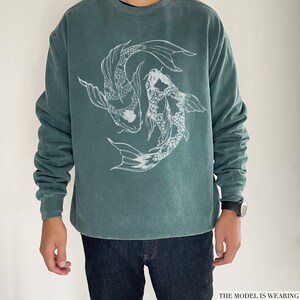 May include: A teal green sweatshirt featuring a white graphic of two koi fish swimming in a circle. The model is wearing one size up from their usual size.
