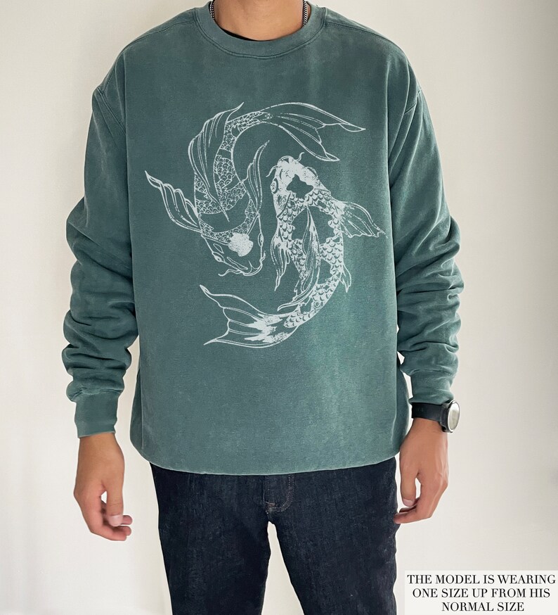 May include: A teal green sweatshirt featuring a white graphic of two koi fish swimming in a circle. The model is wearing one size up from their usual size.