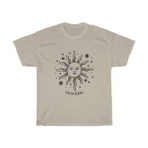 May include: A beige t-shirt with a black graphic of a sun with a face and stars around it. The text "LE SOLEIL" is below the graphic.