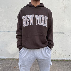 May include: A brown hoodie with a white outline of the words "NEW YORK" in a beige color. The model is wearing the hoodie one size up from their normal size.