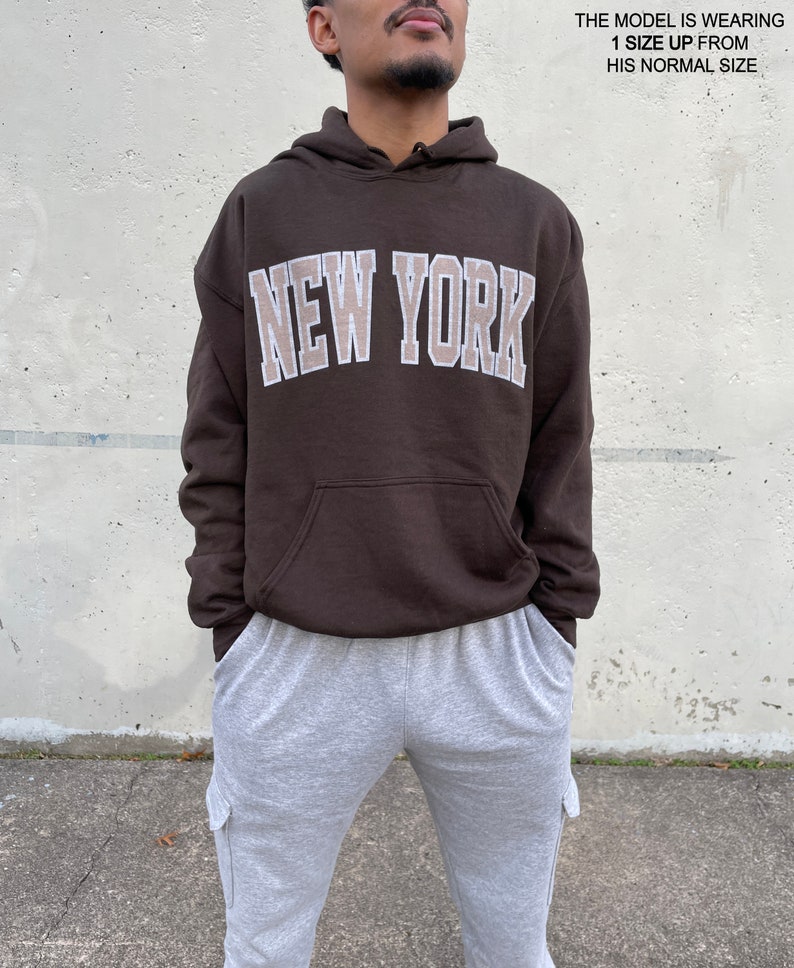 May include: A brown hoodie with a white outline of the words "NEW YORK" in a beige color. The model is wearing the hoodie one size up from their normal size.