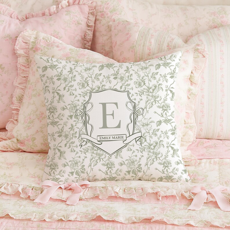 May include: A floral patterned cushion with a grey monogrammed crest featuring the letter "E" and the name "Emily Marie". The cushion is surrounded by other cushions and bedding in shades of pink and white.