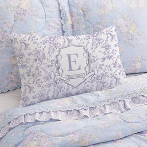May include: A light blue and white floral patterned pillow with a lavender monogrammed "E" in a shield design. The pillow is part of a bedding set with a matching comforter and pillowcases. The comforter and pillowcases have a lace trim.