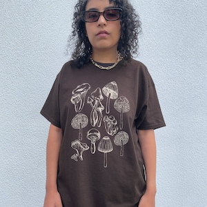 May include: A brown t-shirt with a white outline drawing of various mushrooms. The shirt has a relaxed fit and short sleeves.