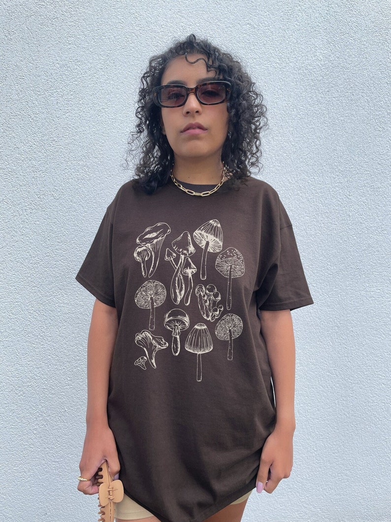May include: A brown t-shirt with a white outline drawing of various mushrooms. The shirt has a relaxed fit and short sleeves.