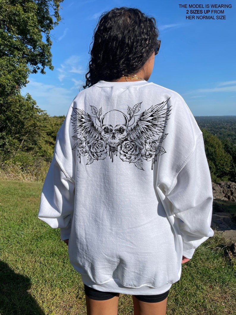 May include: White sweatshirt with a black skull with wings and roses graphic on the back. The model is wearing the sweatshirt two sizes up from her normal size.