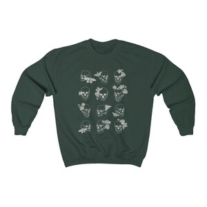 K&ouml;nnte beinhalten: Ein dunkelgr&uuml;ner Pullover mit einem wei&szlig;en Druck von mehreren Sch&auml;deln mit floralen und Schmetterlings-Akzenten.