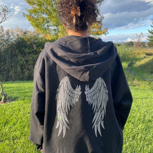 May include: Black hooded sweatshirt with a large rhinestone design of angel wings on the back. The model is wearing the sweatshirt two sizes up from her normal size.