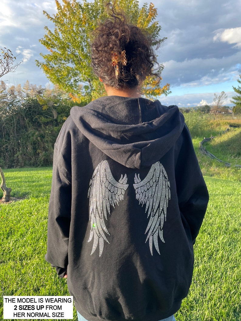 May include: Black hooded sweatshirt with a large rhinestone design of angel wings on the back. The model is wearing the sweatshirt two sizes up from her normal size.