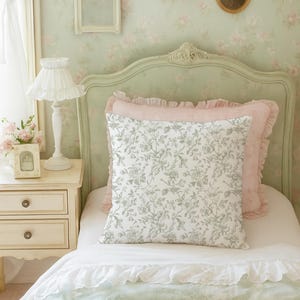 May include: A bedroom scene with a light green headboard and floral wallpaper. A bed is made with a white comforter, a floral patterned pillow, and a pink ruffled pillow. A white nightstand with a lamp and framed photo sits beside the bed.