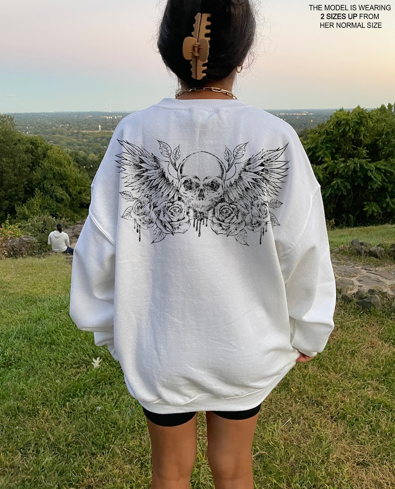 May include: White oversized sweatshirt with a black graphic print of a skull with wings and roses. The model is wearing the sweatshirt two sizes up from her normal size.