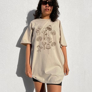 May include: A beige oversized t-shirt with a brown mushroom graphic print. The shirt has short sleeves and a crew neck.