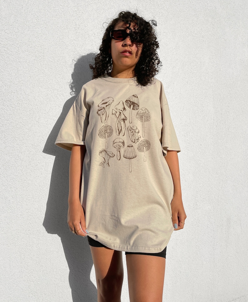 May include: A beige oversized t-shirt with a brown mushroom graphic print. The shirt has short sleeves and a crew neck.