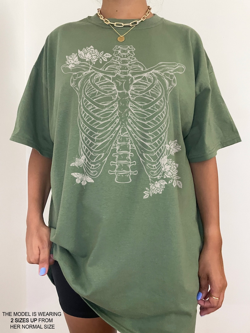 K&ouml;nnte beinhalten: Ein gr&uuml;nes Oversize-T-Shirt mit einem wei&szlig;en Aufdruck eines Brustkorbs und Blumen. Das Model tr&auml;gt das Shirt zwei Gr&ouml;&szlig;en gr&ouml;&szlig;er als seine normale Gr&ouml;&szlig;e.