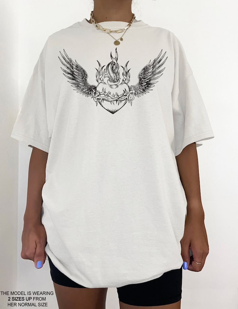 May include: White oversized t-shirt featuring a black graphic of a heart with wings and flames. The heart is surrounded by thorns.