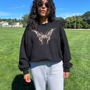 Pode incluir: Camisola de moletom preta com um gr&aacute;fico de borboleta dourada. A modelo est&aacute; usando um tamanho maior do que o seu tamanho normal.