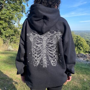 May include: Black hoodie with a white skeleton ribcage graphic on the back. The model is wearing the hoodie two sizes up from their normal size.
