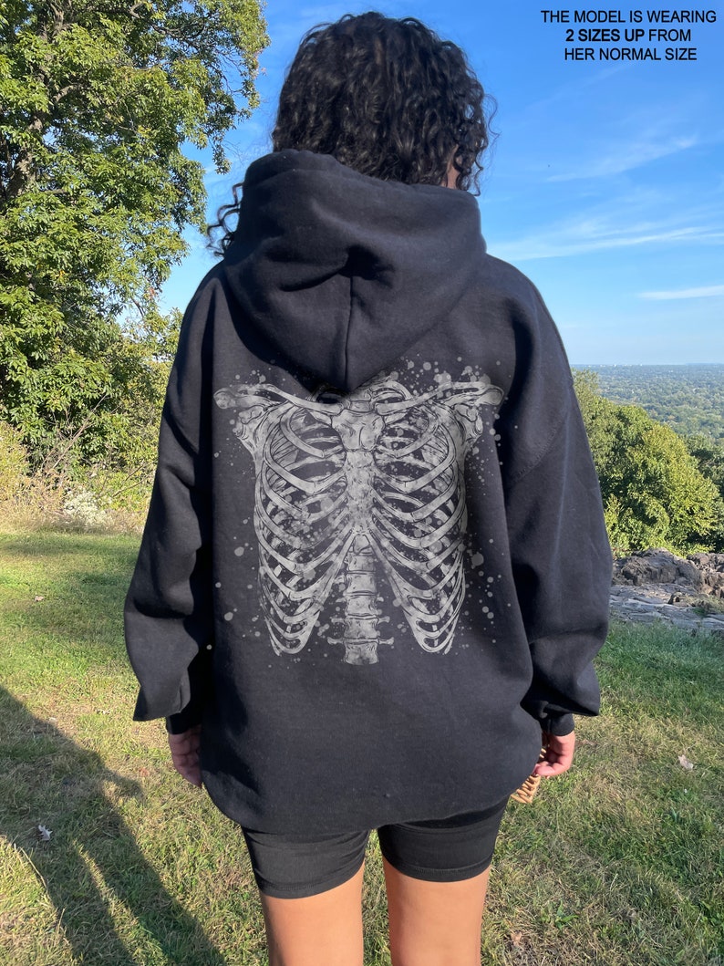 May include: Black hoodie with a white skeleton ribcage graphic on the back. The model is wearing the hoodie two sizes up from their normal size.