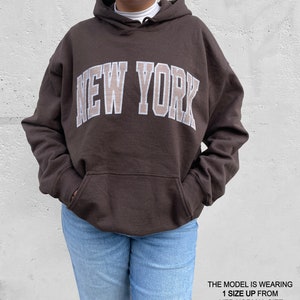 May include: A brown hoodie with a large white "NEW YORK" graphic on the front. The model is wearing a size up from their normal size.