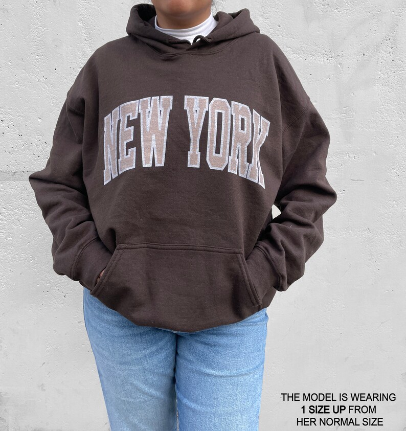 May include: A brown hoodie with a large white "NEW YORK" graphic on the front. The model is wearing a size up from their normal size.