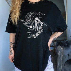 Puede incluir: Camiseta negra de gran tama&ntilde;o con un gr&aacute;fico blanco de dos peces koi nadando en c&iacute;rculo. El texto "Babygirl" es visible en un collar de cadena dorada.