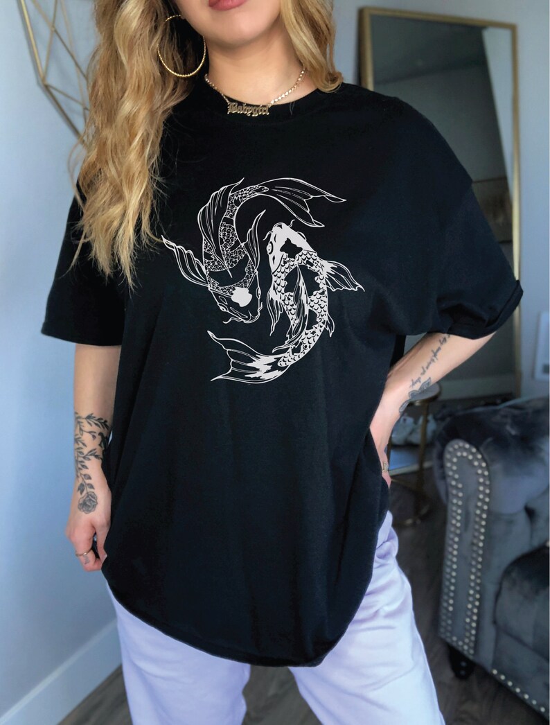 Puede incluir: Camiseta negra de gran tama&ntilde;o con un gr&aacute;fico blanco de dos peces koi nadando en c&iacute;rculo. El texto "Babygirl" es visible en un collar de cadena dorada.