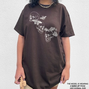 Puede incluir: Una camiseta marr&oacute;n con un gr&aacute;fico de tres calaveras blancas, una mariposa y flores. La modelo lleva la camiseta dos tallas m&aacute;s grande que su talla normal.