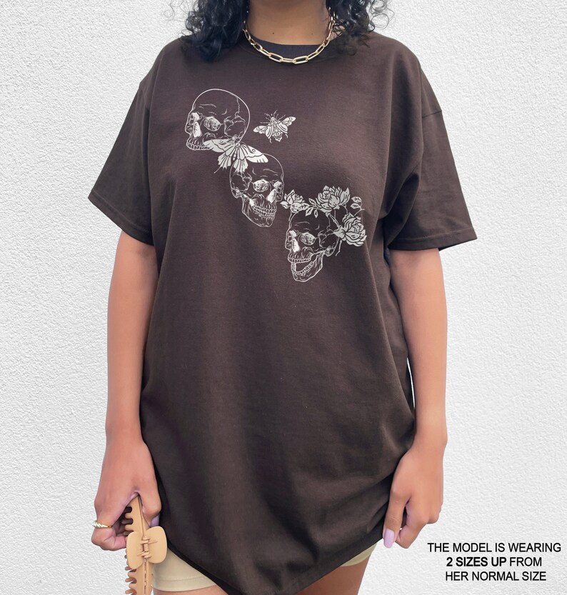 Puede incluir: Una camiseta marr&oacute;n con un gr&aacute;fico de tres calaveras blancas, una mariposa y flores. La modelo lleva la camiseta dos tallas m&aacute;s grande que su talla normal.