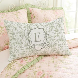 May include: A rectangular pillow with a floral pattern in shades of white and green, featuring a grey crest with the letter "E" and the name "Emily Marie". The pillow is on a bed with pink floral bedding.