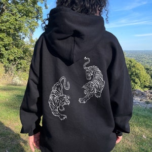 May include: Black hoodie with a white graphic of two tigers facing each other. The model is wearing the hoodie one size up from her usual size.