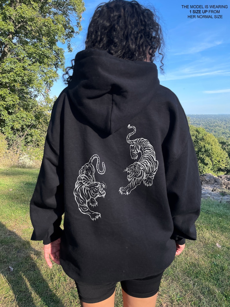 May include: Black hoodie with a white graphic of two tigers facing each other. The model is wearing the hoodie one size up from her usual size.