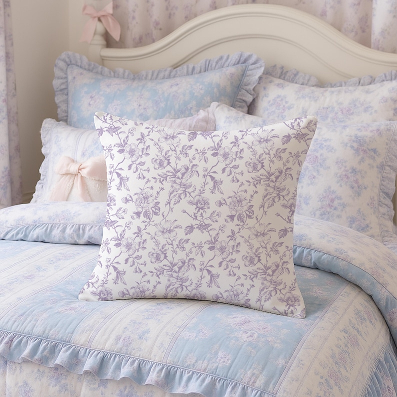 May include: A square decorative pillow with a white background and a purple floral pattern. The pillow is on a bed with a light blue and white floral patterned quilt and matching pillows. The bed has a white headboard and is decorated with a pink bow.