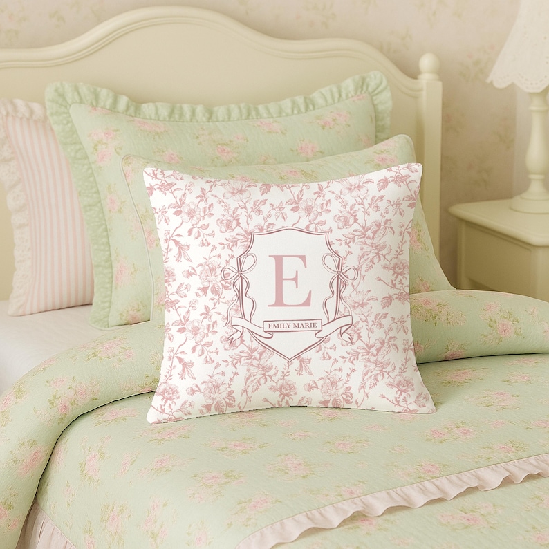 May include: A bed set with a floral pattern in shades of pink and green. The set includes a pillow with the letter "E" and the name "Emily Marie" in a decorative frame. The bedding has a ruffled edge.