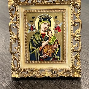 May include: A small rectangular religious icon featuring a depiction of the Virgin Mary holding the Christ child, framed in an ornate gold-colored metal border with an off-white inner trim. The figures are adorned with crowns and halos.