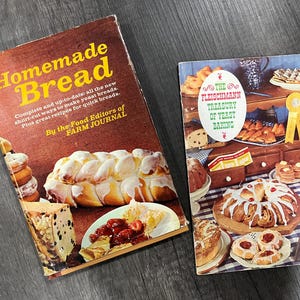 May include: Two vintage cookbooks on a wooden surface. The book on the left is titled "Homemade Bread" with yellow text. The book on the right is titled "The Fleischmann Treasury of Yeast Baking". Both books feature images of baked goods.