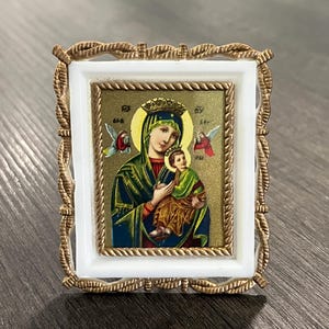 May include: A small rectangular religious icon featuring a depiction of the Virgin Mary holding the Christ child, set against a gold background with two angels. The image is encased in a white frame with an ornate, textured gold-tone metal border.