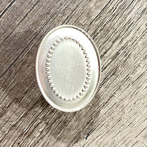 May include: A silver-toned oval pendant featuring a smooth, polished center surface surrounded by a delicate, raised beaded border. The piece has a clean, metallic finish and a simple, symmetrical design that highlights the contrast between the smooth inner oval and the textured, circular bead detailing along the perimeter.
