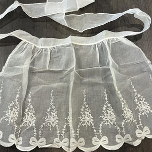 May include: A white sheer waist apron featuring a gathered skirt with a scalloped hemline. The fabric is decorated with white floral embroidery and small bow motifs along the bottom edge. It includes a long tie for securing around the waist.